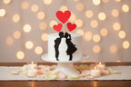Small white cake with black silhouettes of a couple and red heart-shaped toppers on a white background.